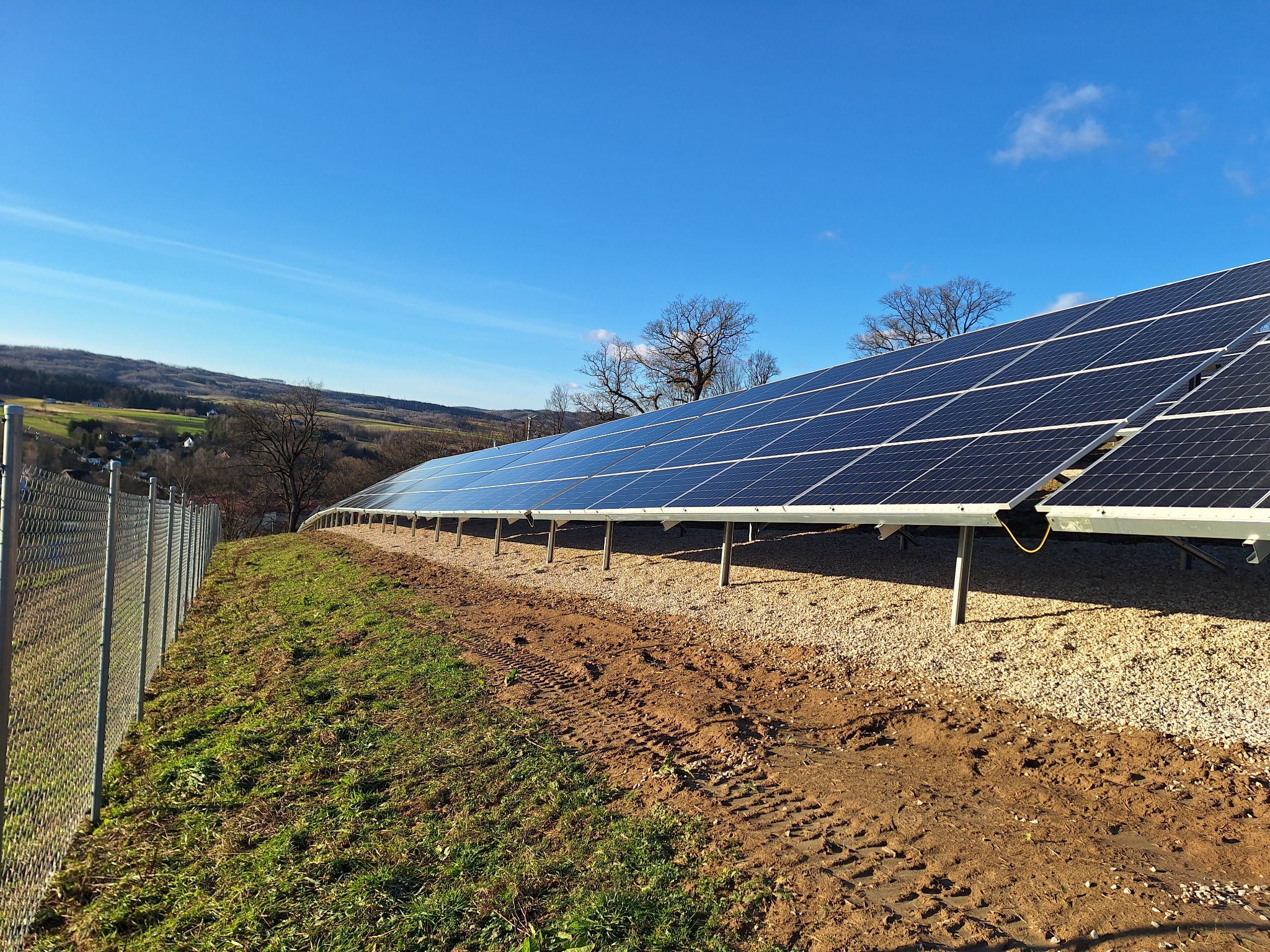 Rząd paneli fotowoltaicznych na żwirowym podłożu, widok z dołu, ślady opon na ziemi, w tle drzewa i błękitne niebo.