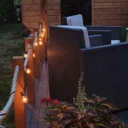 Oświetlony taras z drewnianą sauną w tle, rattanowe meble ogrodowe i dekoracyjne lampki sznurkowe. Donica z kolorowymi liśćmi roślin w pierwszym planie.