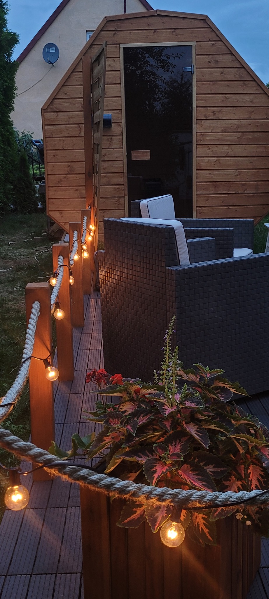 Oświetlony taras z drewnianą sauną w tle, rattanowe meble ogrodowe i dekoracyjne lampki sznurkowe. Donica z kolorowymi liśćmi roślin w pierwszym planie.