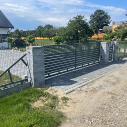 Kuźnia Stali ROKSANA ZADWORNA - Nowoczesna, pozioma brama wjazdowa z szarych paneli, osadzona na słupkach z szarej kostki, prowadząca na posesję z widocznym domem i ogrodem w tle.