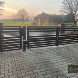 Nowoczesna, pozioma brama wjazdowa z automatyką, wykonana z ciemnego metalu, na tle kostki brukowej i krajobrazu z budynkiem w oddali, widoczny mechanizm otwierający.