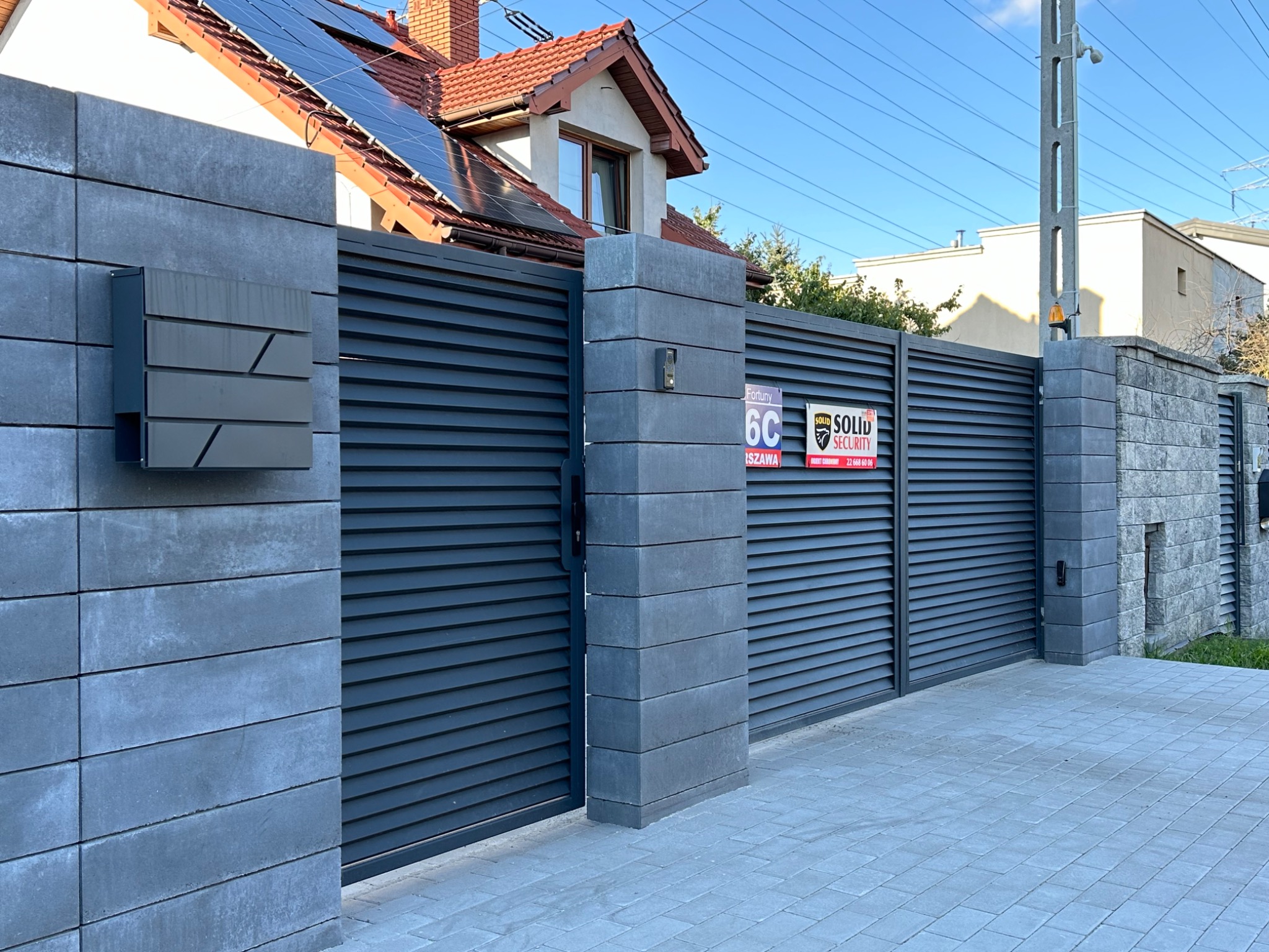 Nowoczesna posesja z szarą, poziomą bramą lamelową, murowanymi słupkami z betonu architektonicznego i skrzynką na listy o geometrycznym designie.