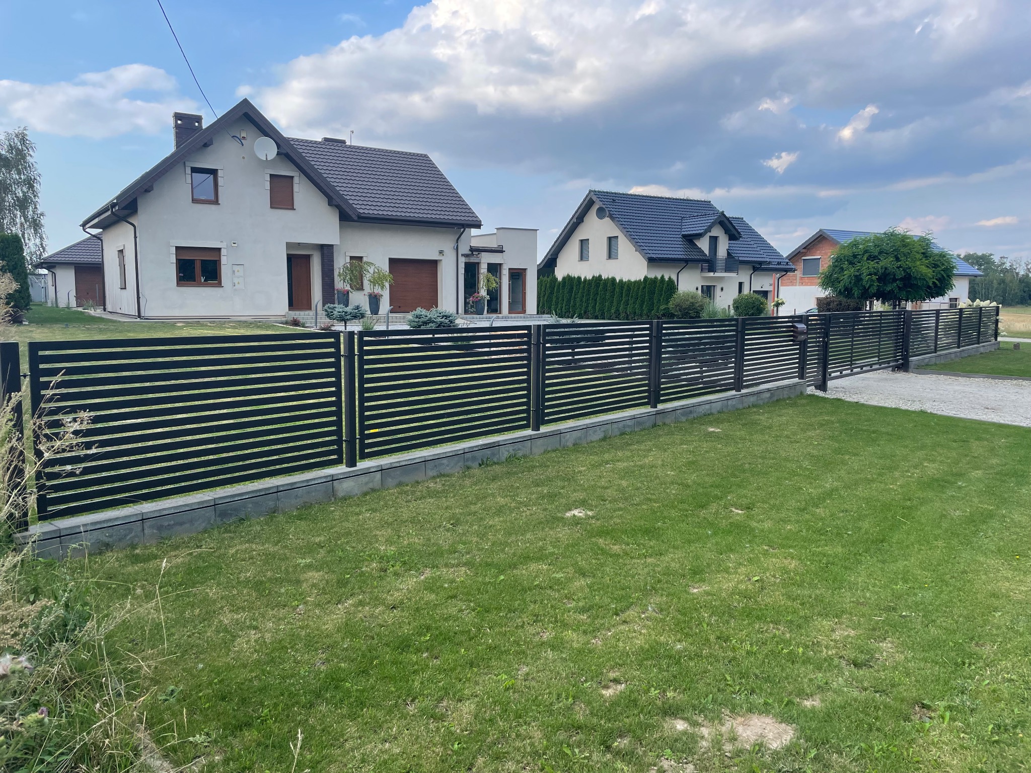 Nowoczesne, czarne ogrodzenie metalowe z poziomymi panelami na podmurówce z szarej kostki, oddzielające posesję z domami jednorodzinnymi od drogi.