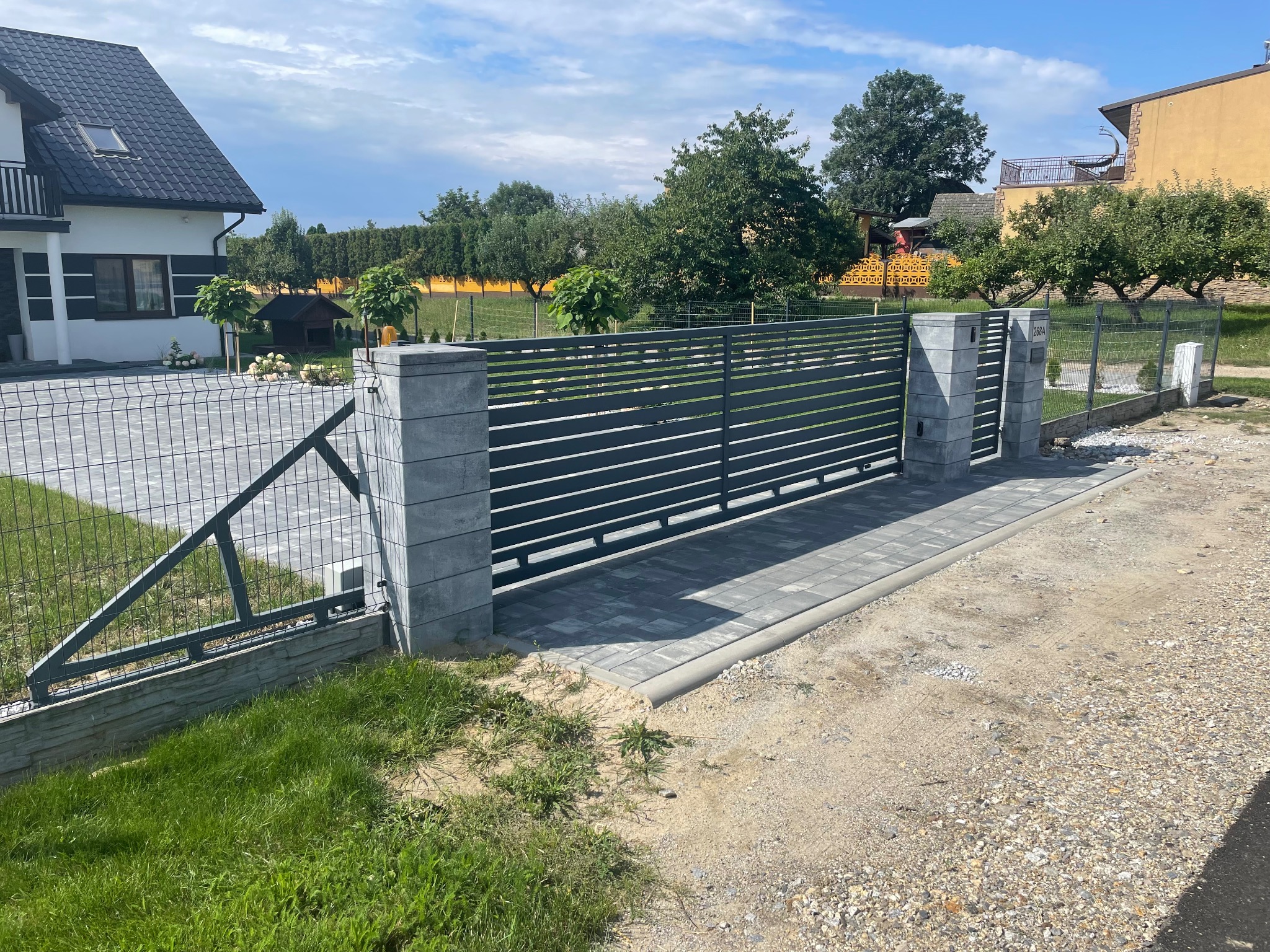 Nowoczesna, pozioma brama wjazdowa z szarych paneli, osadzona na słupkach z szarej kostki, prowadząca na posesję z widocznym domem i ogrodem w tle.