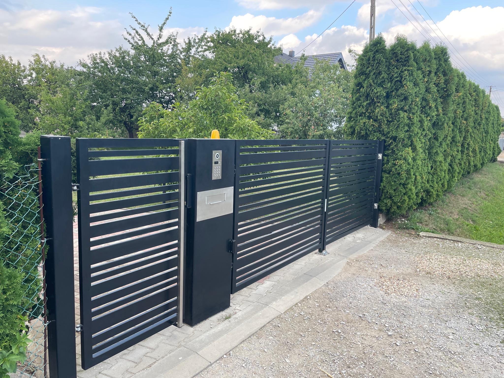 Nowoczesna brama wjazdowa przesuwna z poziomych profili, zintegrowana ze skrzynką na listy i panelem domofonu, na tle zieleni i żwirowego podjazdu.