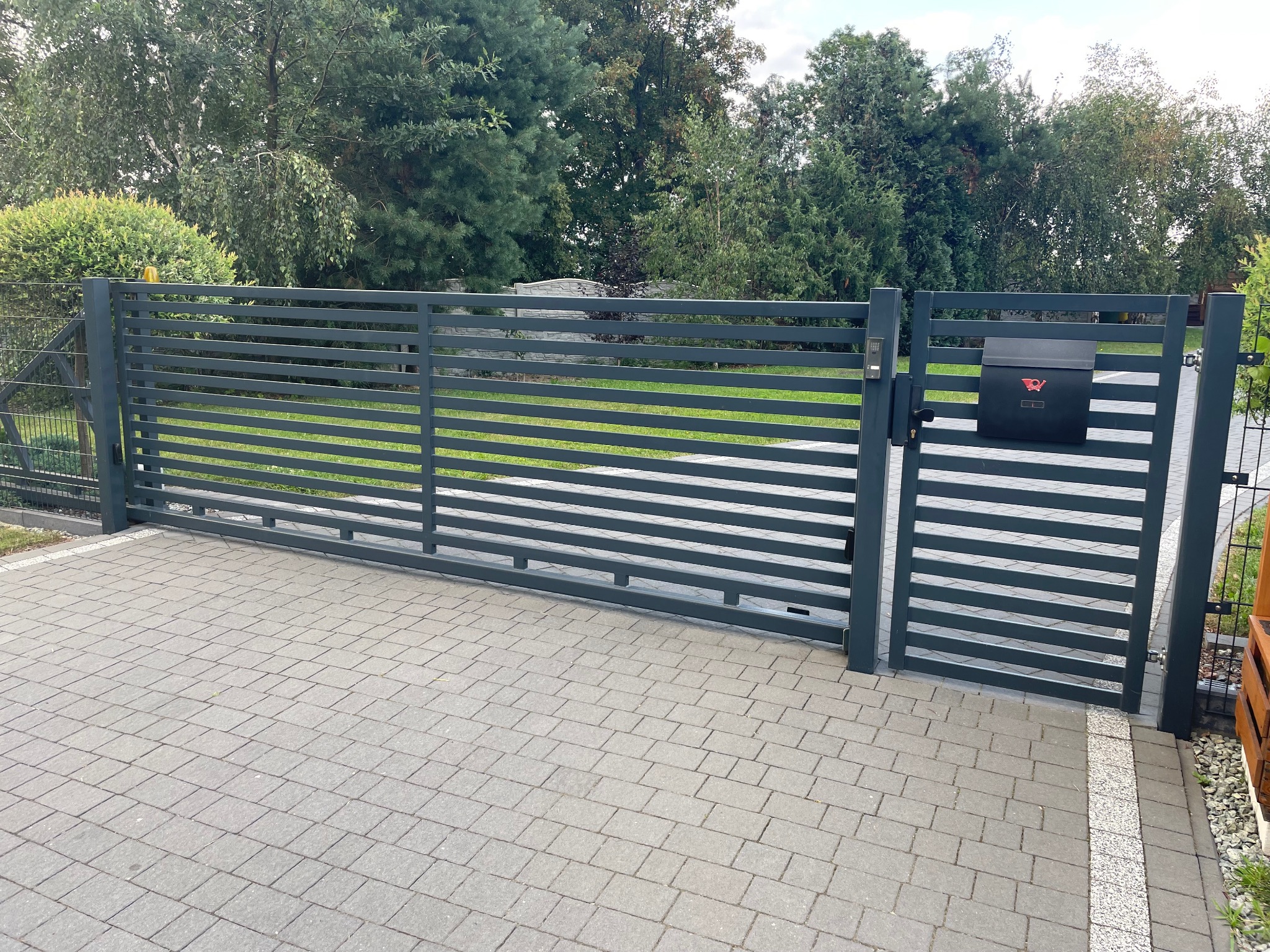 Nowoczesna, pozioma brama wjazdowa i furtka w kolorze antracytowym, z zamontowaną skrzynką pocztową z symbolem poczty, na tle zielonego trawnika i drzew.