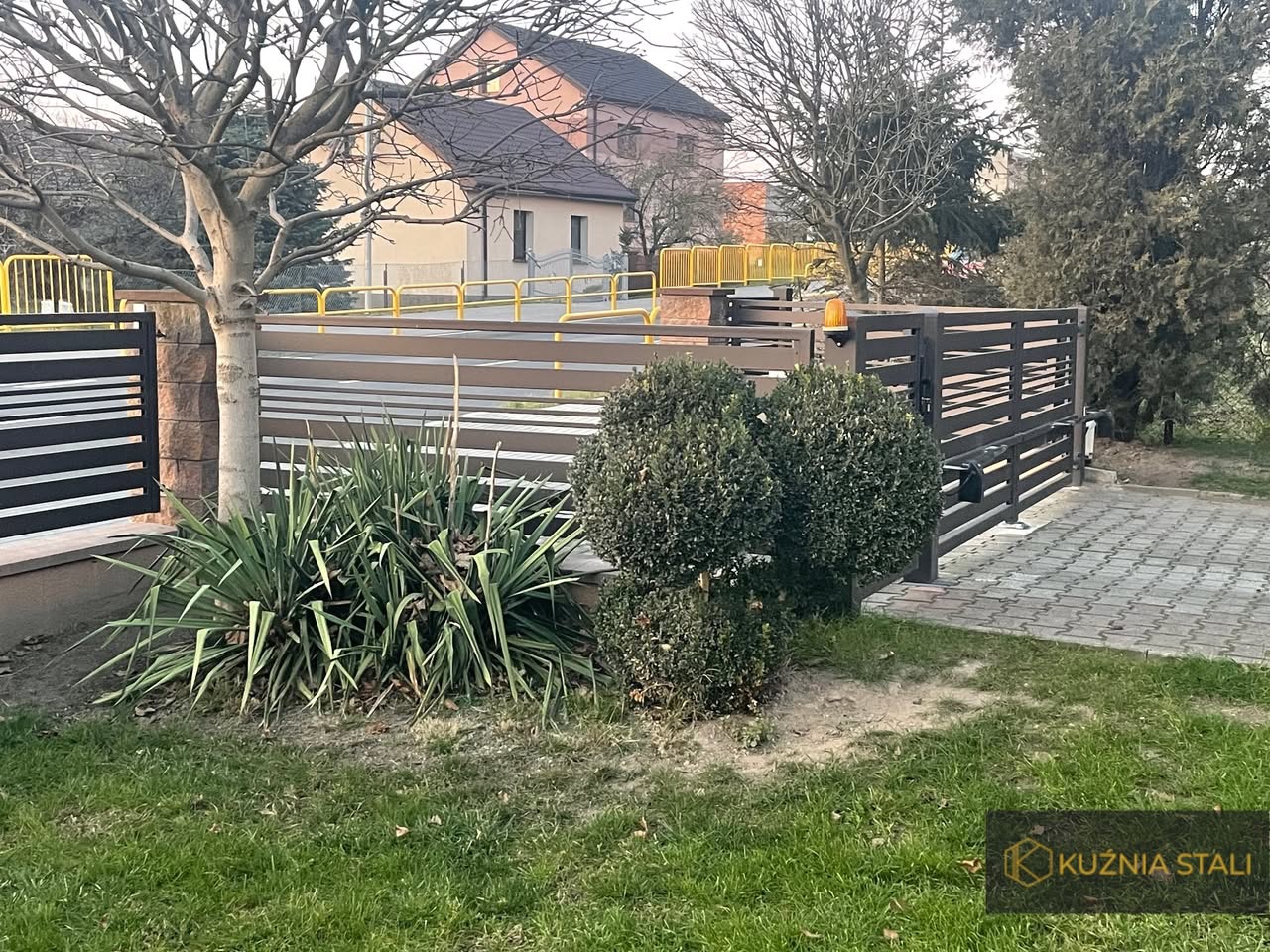 Nowoczesna brama wjazdowa przesuwna z automatyką, widoczna z zewnątrz posesji, z elementami drewnopodobnymi i szarymi panelami, otoczona zielenią i kostką brukową.