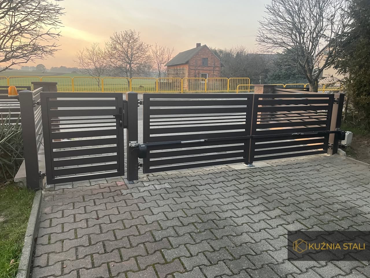 Nowoczesna, pozioma brama wjazdowa z automatyką, wykonana z ciemnego metalu, na tle kostki brukowej i krajobrazu z budynkiem w oddali, widoczny mechanizm otwierający.