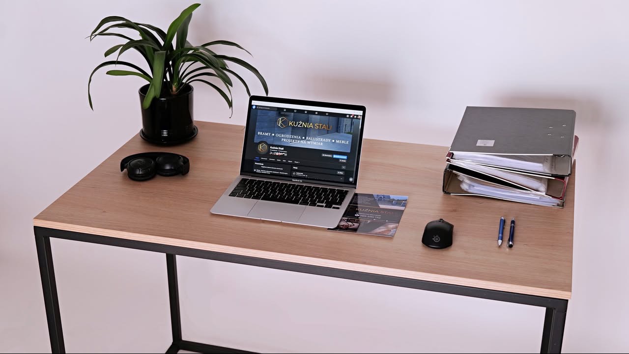 Biurko z laptopem wyświetlającym stronę Kuźnia Stali, obok leżą słuchawki, myszka, dwa długopisy, ulotka firmy i segregator z dokumentami, a w tle roślina doniczkowa.