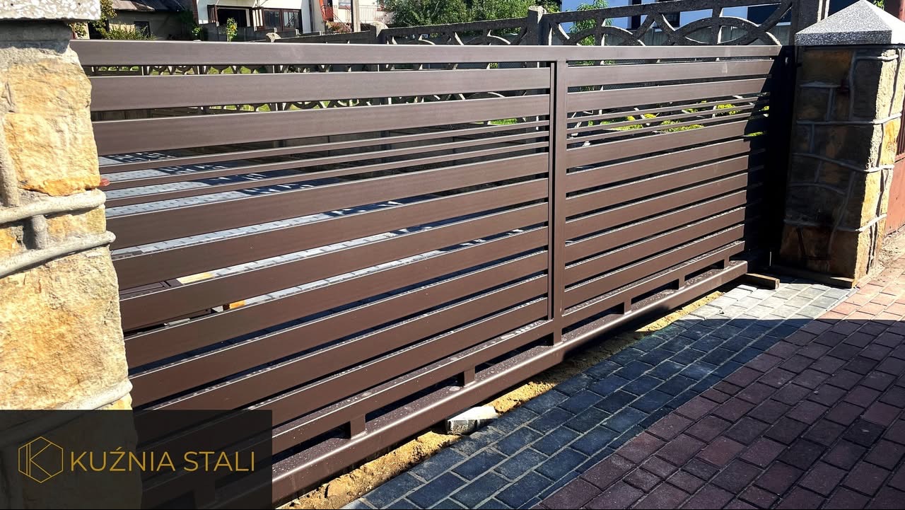 Przesuwna brama posesyjna z poziomych profili w kolorze brązowym, osadzona między kamiennymi słupkami, widoczny fragment kostki brukowej i logo firmy Kuźnia Stali.