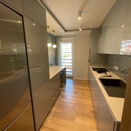 Furniture Mateusz Zuber - Wąska kuchnia z szarymi, lakierowanymi frontami, wyspą z białym blatem, drewnianą podłogą i oknem z widokiem na balkon.
