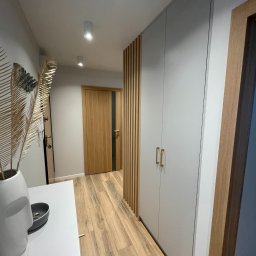 Furniture Mateusz Zuber - Korytarz z zabudowaną szafą w kolorze szarym z złotymi uchwytami, drewnianą podłogą i dekoracyjnymi lamelami na ścianie, widok z perspektywy wejścia, z elementami dekoracyjnymi na pierwszym planie.
