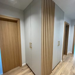 Furniture Mateusz Zuber - Szara szafa wnękowa z drewnianymi lamelami dekoracyjnymi i złotymi uchwytami, widoczna w korytarzu z drewnianą podłogą i drzwiami.