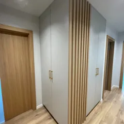 Szara szafa wnękowa z drewnianymi lamelami dekoracyjnymi i złotymi uchwytami, widoczna w korytarzu z drewnianą podłogą i drzwiami.