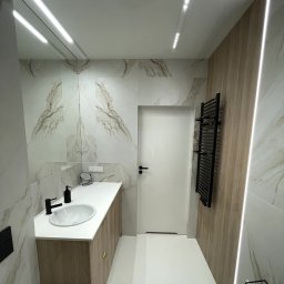 Furniture Mateusz Zuber - Nowoczesna łazienka w Trzebnicy z umywalką nablatową na drewnianej szafce, marmurowe płytki na ścianach, czarny grzejnik drabinkowy i oświetlenie LED.