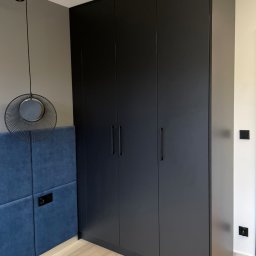 Furniture Mateusz Zuber - Czarna, matowa szafa wnękowa z czterema drzwiami, długie, minimalistyczne uchwyty, widoczny fragment wezgłowia łóżka obitego niebieskim welurem i designerska lampa wisząca.