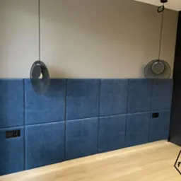Niebieski, tapicerowany panel ścienny w sypialni z dwoma czarnymi gniazdkami elektrycznymi, oświetlony dwoma nowoczesnymi lampami wiszącymi z czarnymi kloszami, z szafą na ubrania w tle.