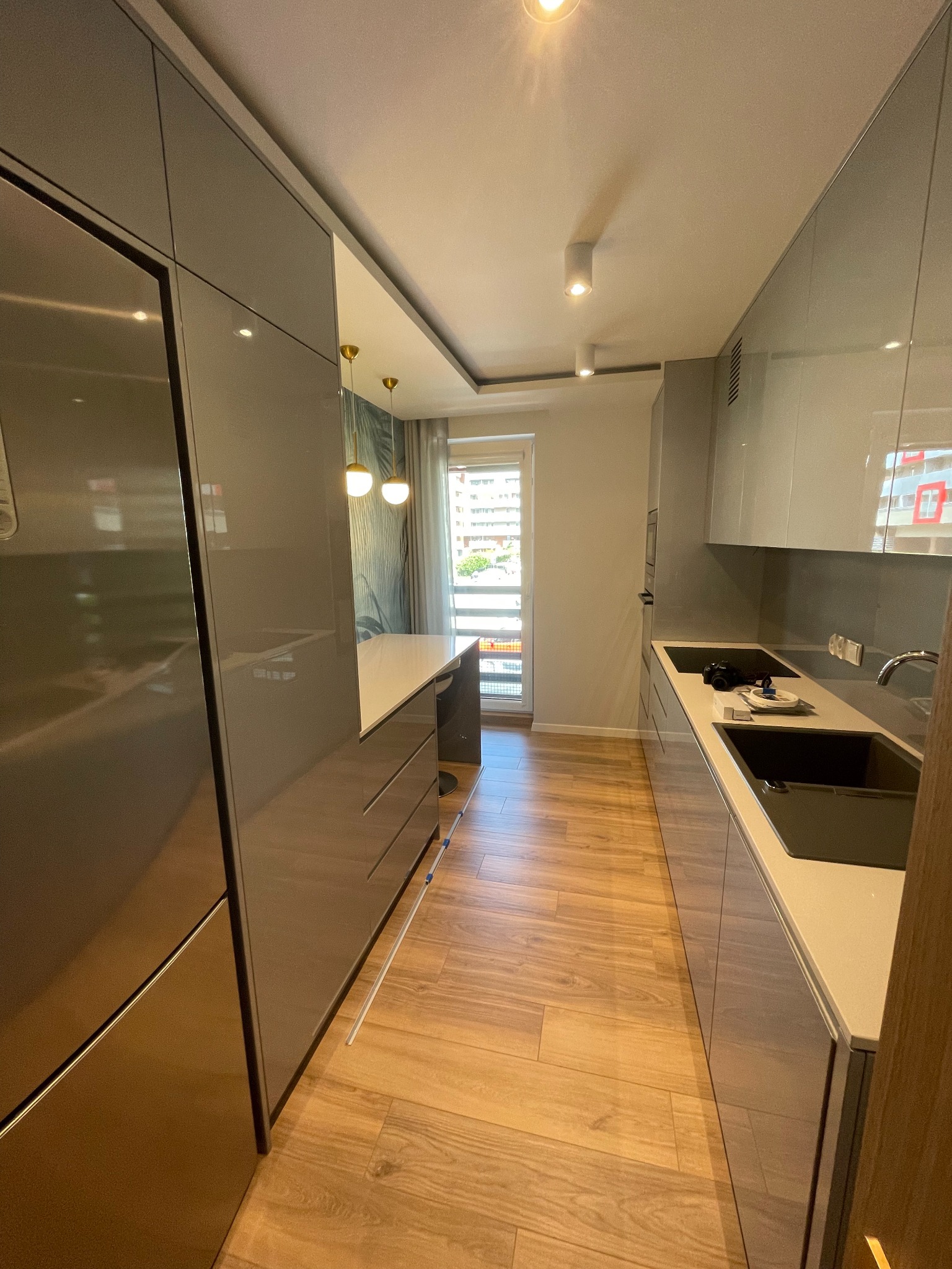 Wąska kuchnia z szarymi, lakierowanymi frontami, wyspą z białym blatem, drewnianą podłogą i oknem z widokiem na balkon.