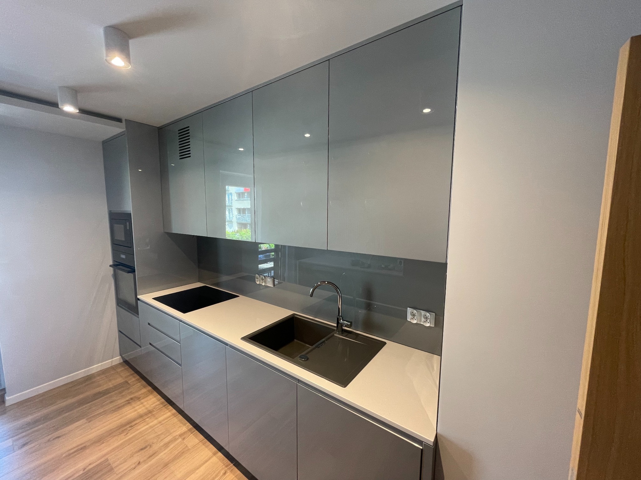 Nowoczesna kuchnia na wymiar w odcieniach szarości z błyszczącymi frontami, czarnym zlewem i płytą indukcyjną, oświetleniem punktowym i drewnianą podłogą.