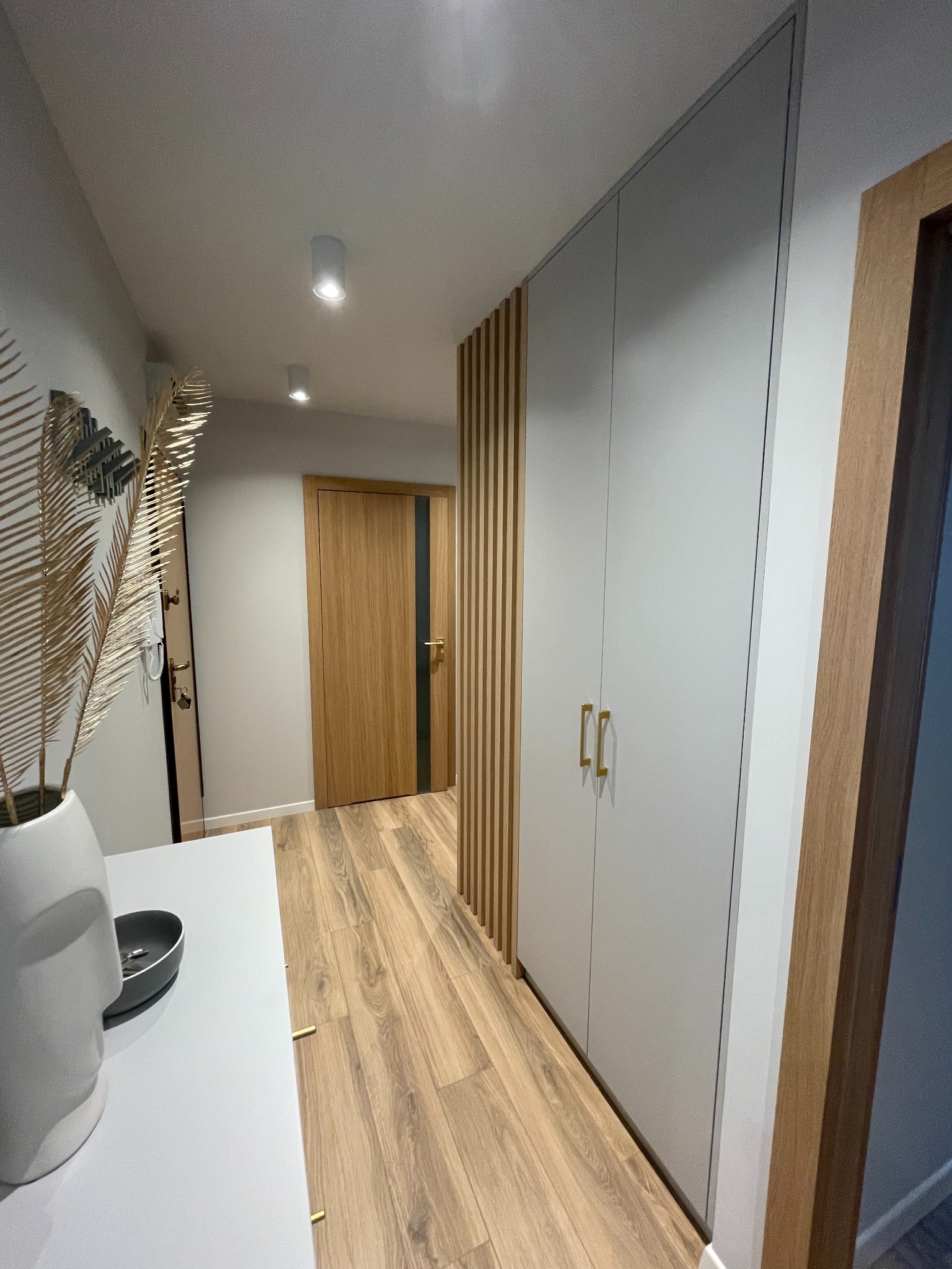 Korytarz z zabudowaną szafą w kolorze szarym z złotymi uchwytami, drewnianą podłogą i dekoracyjnymi lamelami na ścianie, widok z perspektywy wejścia, z elementami dekoracyjnymi na pierwszym planie.