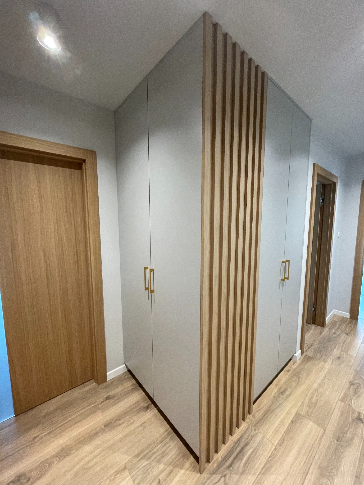 Szara szafa wnękowa z drewnianymi lamelami dekoracyjnymi i złotymi uchwytami, widoczna w korytarzu z drewnianą podłogą i drzwiami.