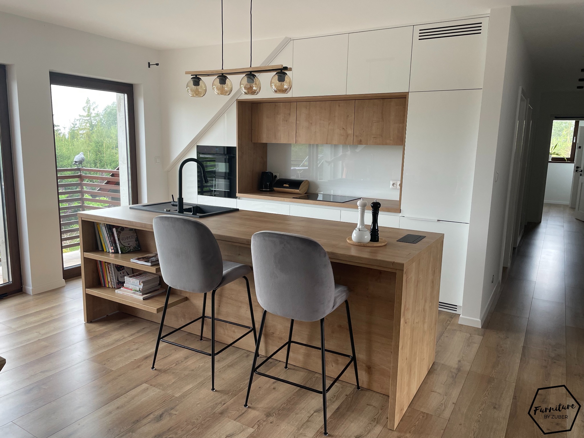 Nowoczesna kuchnia z wyspą, białe lakierowane fronty szafek górnych i drewnopodobne dolne, czarny zlew i bateria, dwie szare welurowe hokery z czarnymi nogami, widok na zewnątrz przez okno balkonowe.