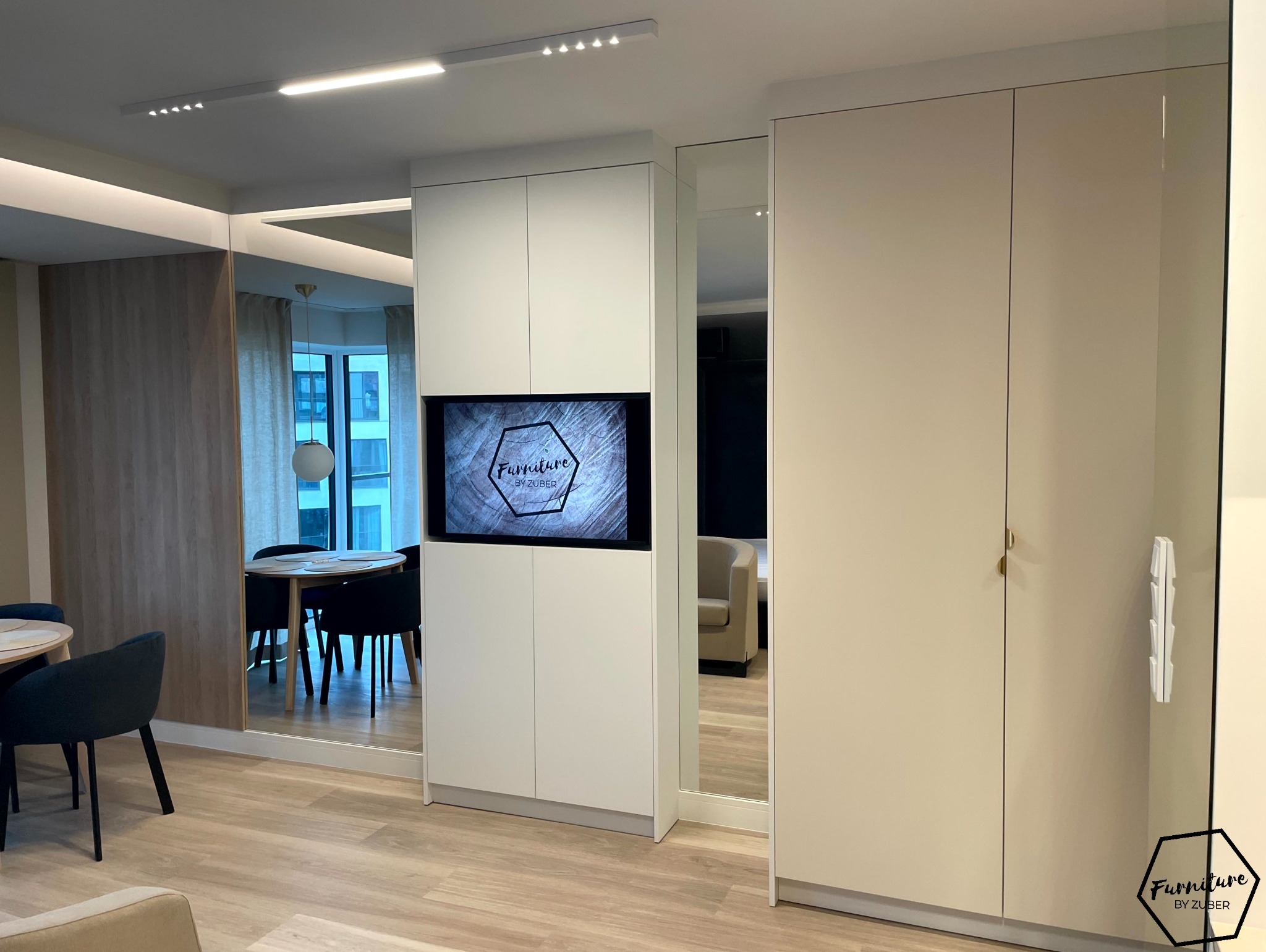 Nowoczesna zabudowa meblowa z białymi szafami i lustrami, wbudowany telewizor z logo firmy meblarskiej, widok na jadalnię i salon w tle.