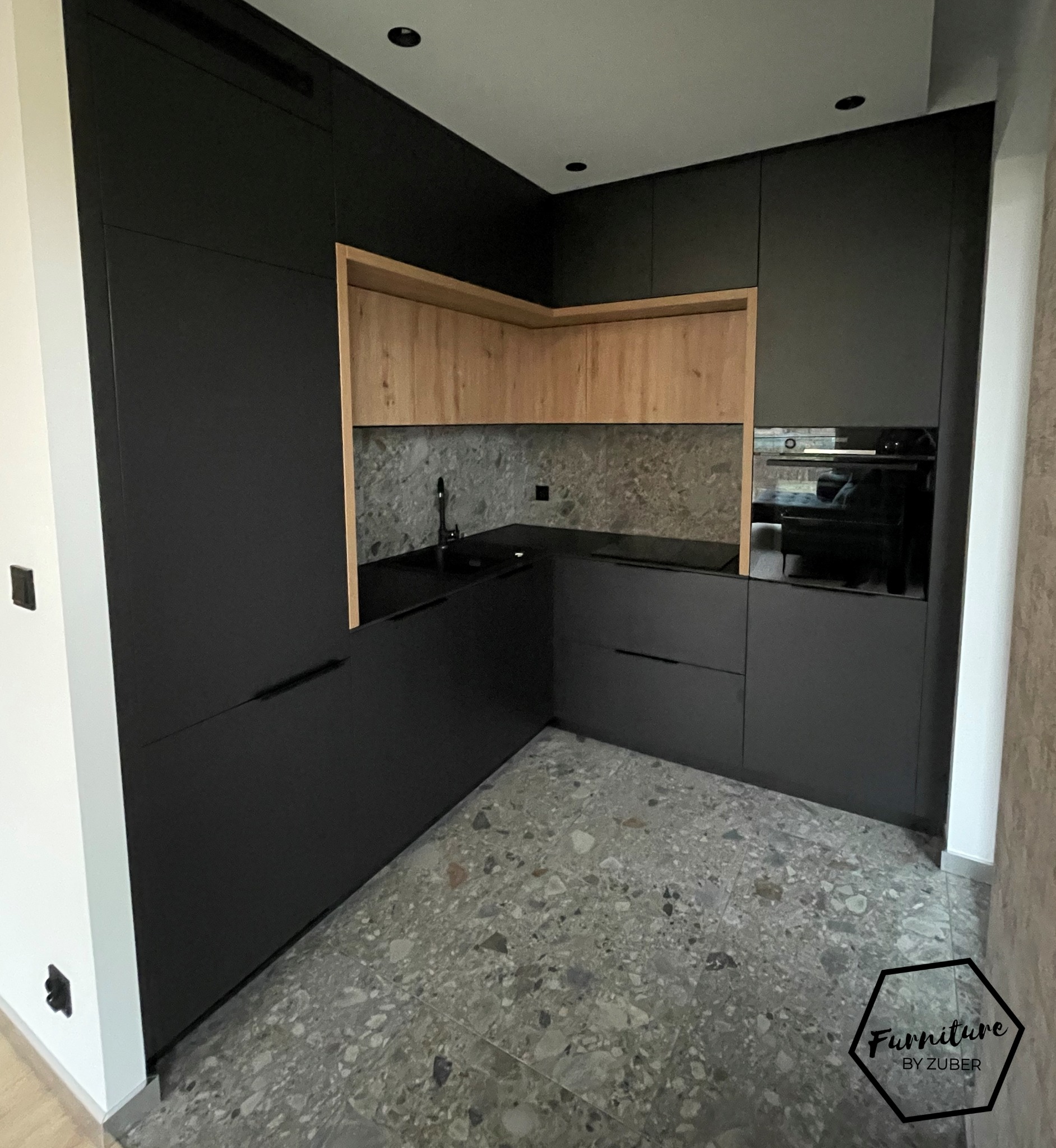Nowoczesna, narożna zabudowa kuchenna z czarnymi, matowymi frontami i drewnianymi szafkami górnymi, uzupełniona blatem w kolorze czerni i szarym backsplashiem, oświetlona punktowymi lampami...