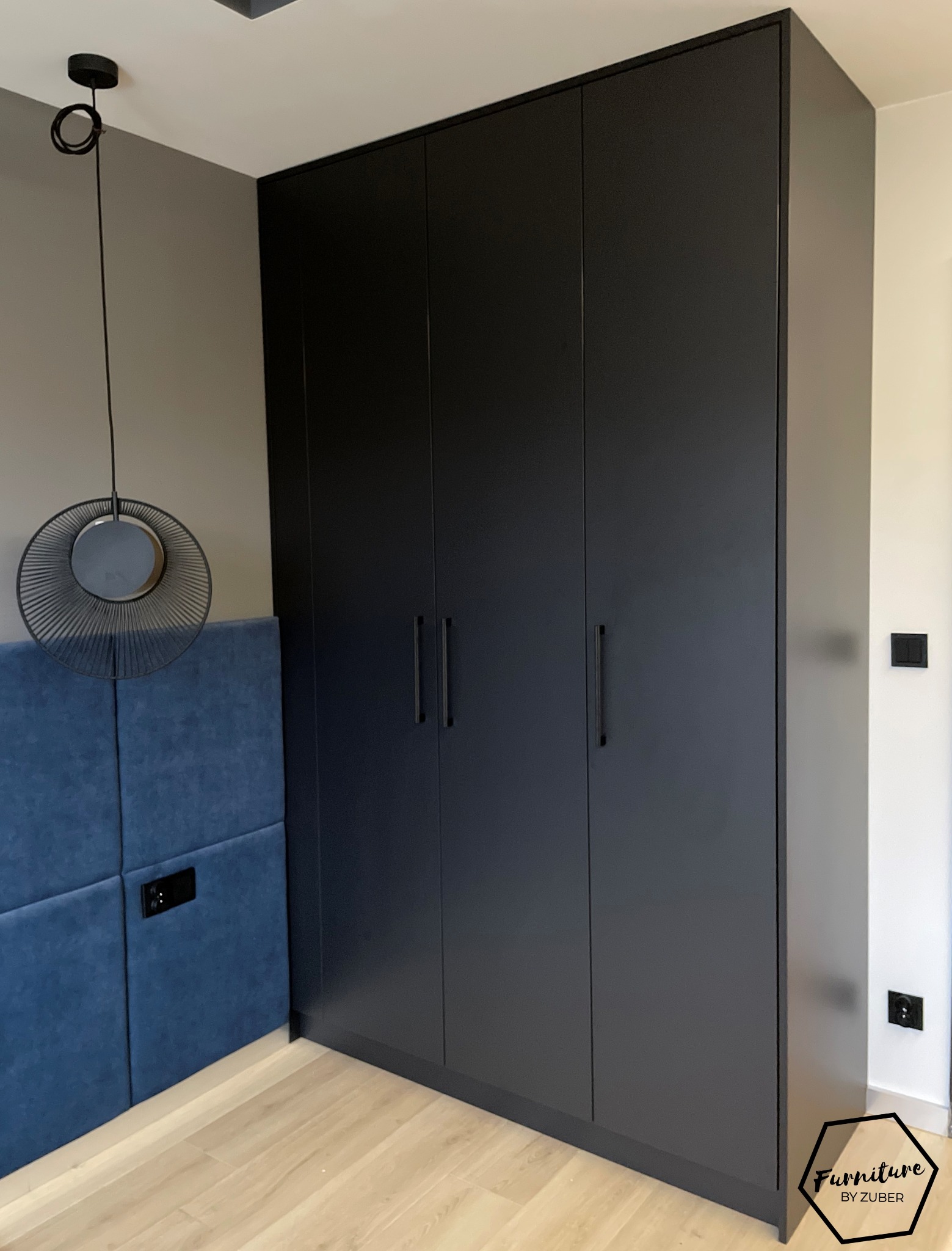 Czarna, matowa szafa wnękowa z czterema drzwiami, długie, minimalistyczne uchwyty, widoczny fragment wezgłowia łóżka obitego niebieskim welurem i designerska lampa wisząca.