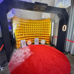 Instalacja z czerwonym dywanem, złotym, dmuchanym tłem, kartami do gry i żetonami kasynowymi, otoczona czarną konstrukcją i światłami LED, z efektem dymu na dywanie.