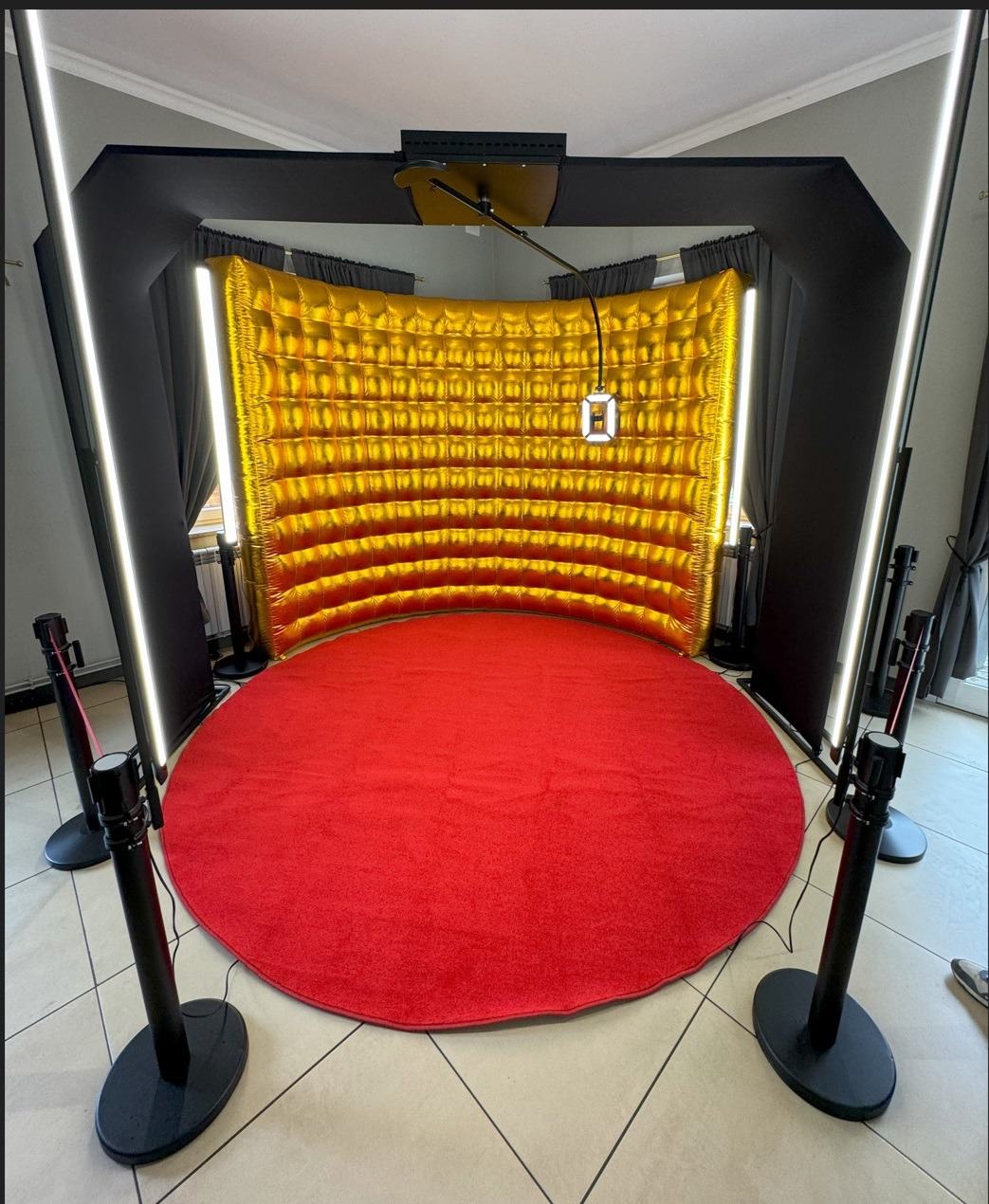 Fotobudka z dmuchanym, złotym tłem, oświetleniem LED i czerwonym dywanem, otoczona barierkami.