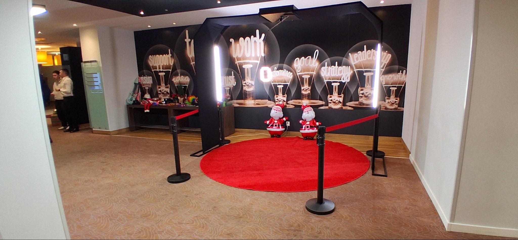 Ustawiona w pomieszczeniu konstrukcja z lampami do zdjęć, czerwony dywan, dwa dmuchane Mikołaje i rekwizyty na stole, tło z motywem żarówek i napisami.