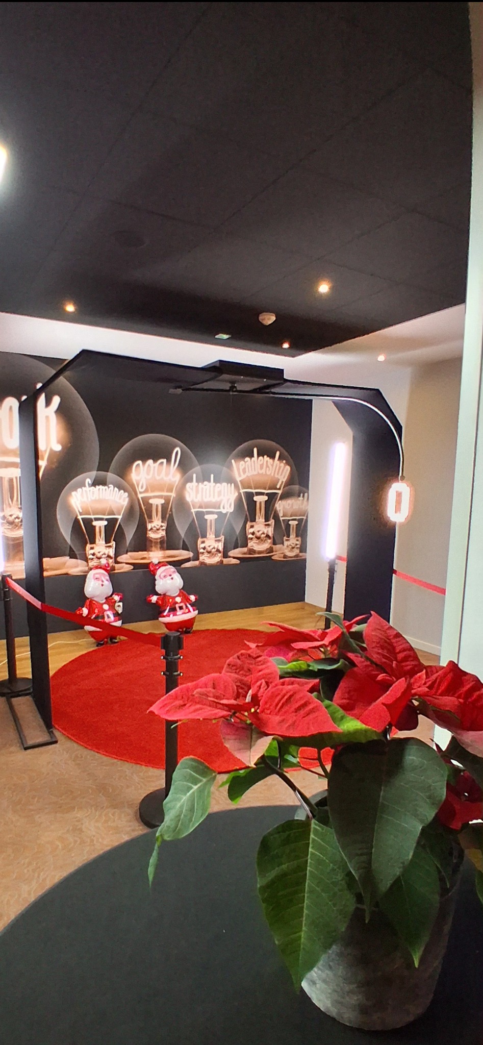 Dekoracja świąteczna z dwoma figurkami Świętego Mikołaja na czerwonym dywanie przed ścianą z grafiką żarówek z napisami 'performance', 'goal', 'strategy', 'leadership', 'growth', oraz poinsecja...