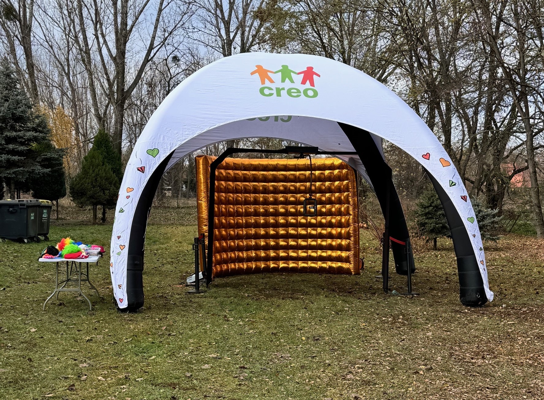 Rozstawiona biała konstrukcja namiotowa z logo firmy i dekoracjami w kształcie serc i samolotów, wewnątrz złota, dmuchana ścianka, obok stolik z kolorowymi perukami i akcesoriami.