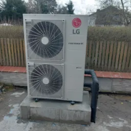 Zewnętrzna jednostka pompy ciepła LG Therma V, zamontowana na betonowym fundamencie przed ogrodzeniem z drewnianych sztachet. Widoczne czarne, izolowane rury przyłączeniowe.