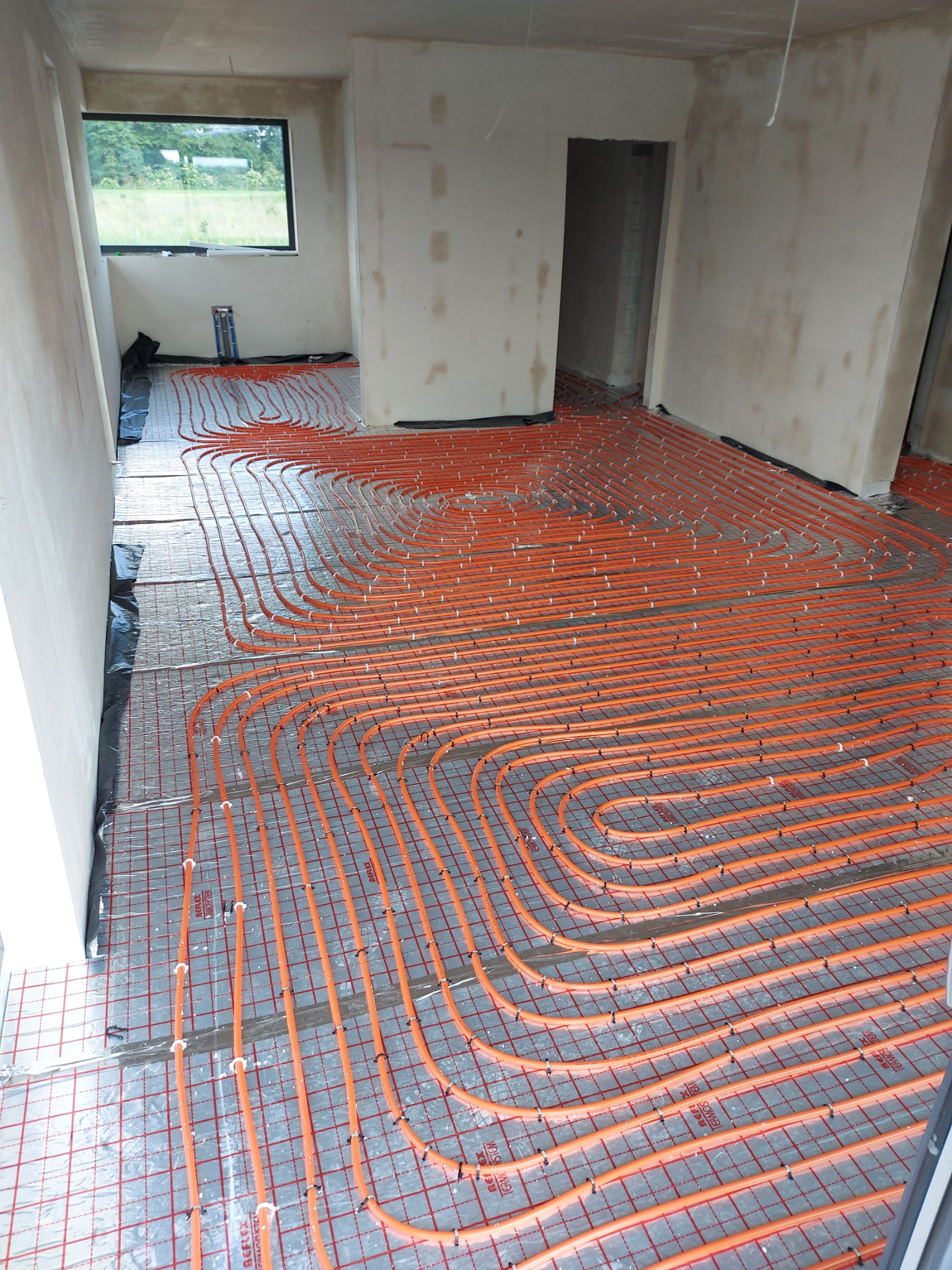 Pomarańczowe rury ogrzewania podłogowego rozłożone na srebrnej macie w surowym wnętrzu, widok z góry na układ pętli przed wylewką.