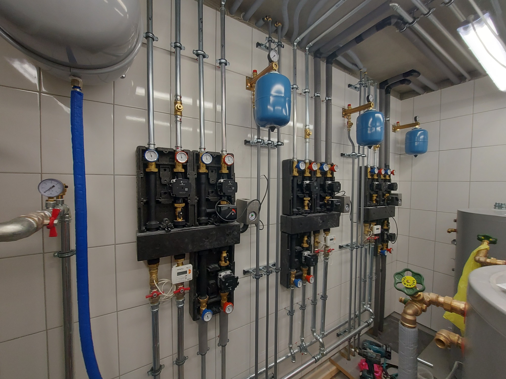 Rozbudowana instalacja hydrauliczna i grzewcza z licznymi zaworami, rurami, manometrami i zbiornikami wyrównawczymi, zamontowana na ścianie z białych płytek ceramicznych w pomieszczeniu technicznym.