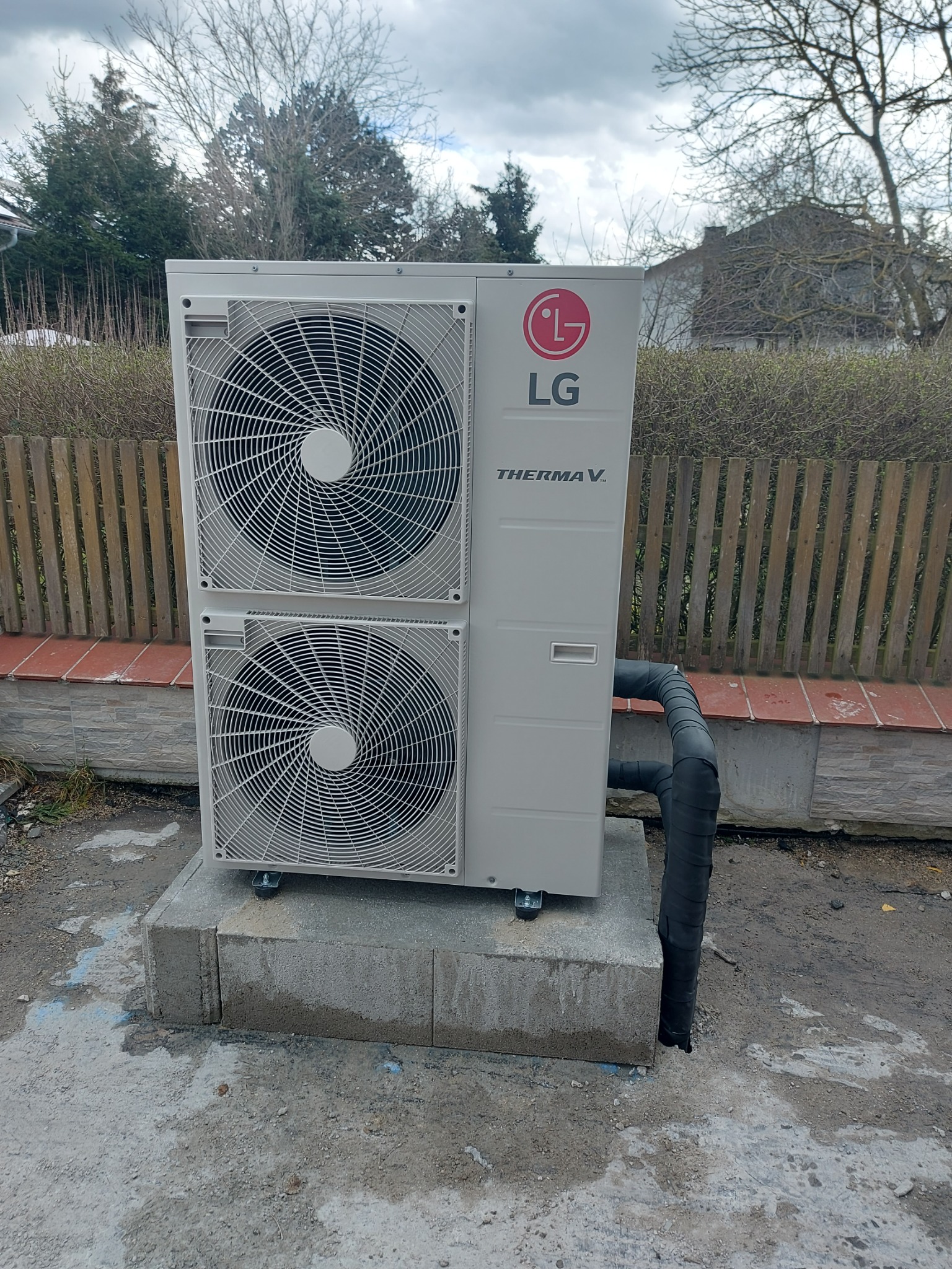 Zewnętrzna jednostka pompy ciepła LG Therma V, zamontowana na betonowym fundamencie przed ogrodzeniem z drewnianych sztachet. Widoczne czarne, izolowane rury przyłączeniowe.