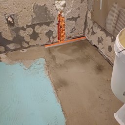 WojtekTom **EleganckieRęce** - Przygotowanie podłoża w łazience: wylewka samopoziomująca częściowo pokryta niebieską hydroizolacją, ściany z resztkami starej glazury, wypełniona pianką montażową szczelina przy podłodze.