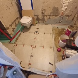 WojtekTom **EleganckieRęce** - Niedokończona łazienka w trakcie układania jasnych, kwadratowych płytek podłogowych z użyciem krzyżyków dystansowych, częściowo otynkowane ściany z widoczną instalacją toalety podtynkowej i pianą...