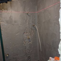 WojtekTom **EleganckieRęce** - Przygotowanie ściany pod płytki w rogu pomieszczenia, widoczna siatka pod tynkiem, poziomica laserowa wyznacza linię, fragment instalacji wodnej, dwa wiadra na podłodze.