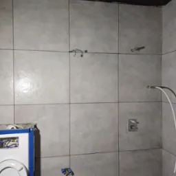 Szare, matowe płytki ścienne w trakcie montażu w łazience, widoczne wystające przewody elektryczne, instalacja wodna i częściowo zamontowana toaleta podtynkowa.