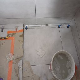 WojtekTom **EleganckieRęce** - Szare płytki na podłodze, w trakcie układania, widoczne narzędzia poziomujące i wiadro z zaprawą, ochraniacz na but.