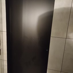 WojtekTom **EleganckieRęce** - Czarne, matowe drzwi wewnętrzne zamontowane w ościeżnicy, widoczny fragment ściany z jasnymi, kwadratowymi płytkami.