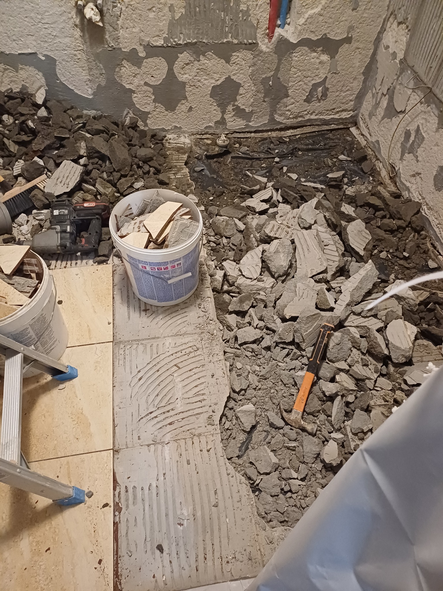 Demontaż starej glazury w łazience, widoczne gruz, resztki kleju na ścianach, wiadro z fragmentami płytek oraz narzędzia: młotek i szlifierka.