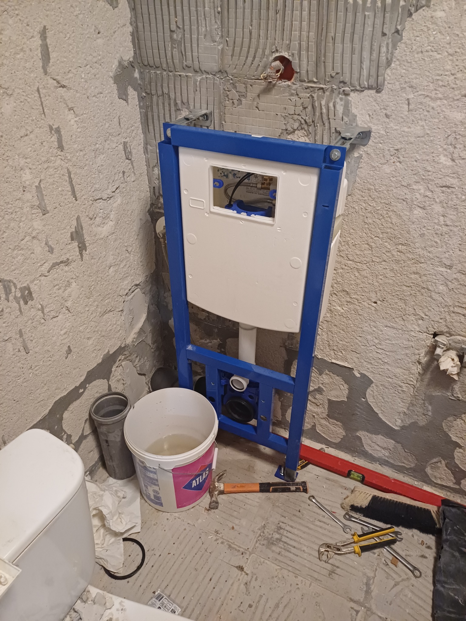 Stelaż podtynkowy WC zamontowany na tle surowej ściany, widoczne narzędzia, wiadro z wodą i fragment toalety, etap prac instalacyjnych.