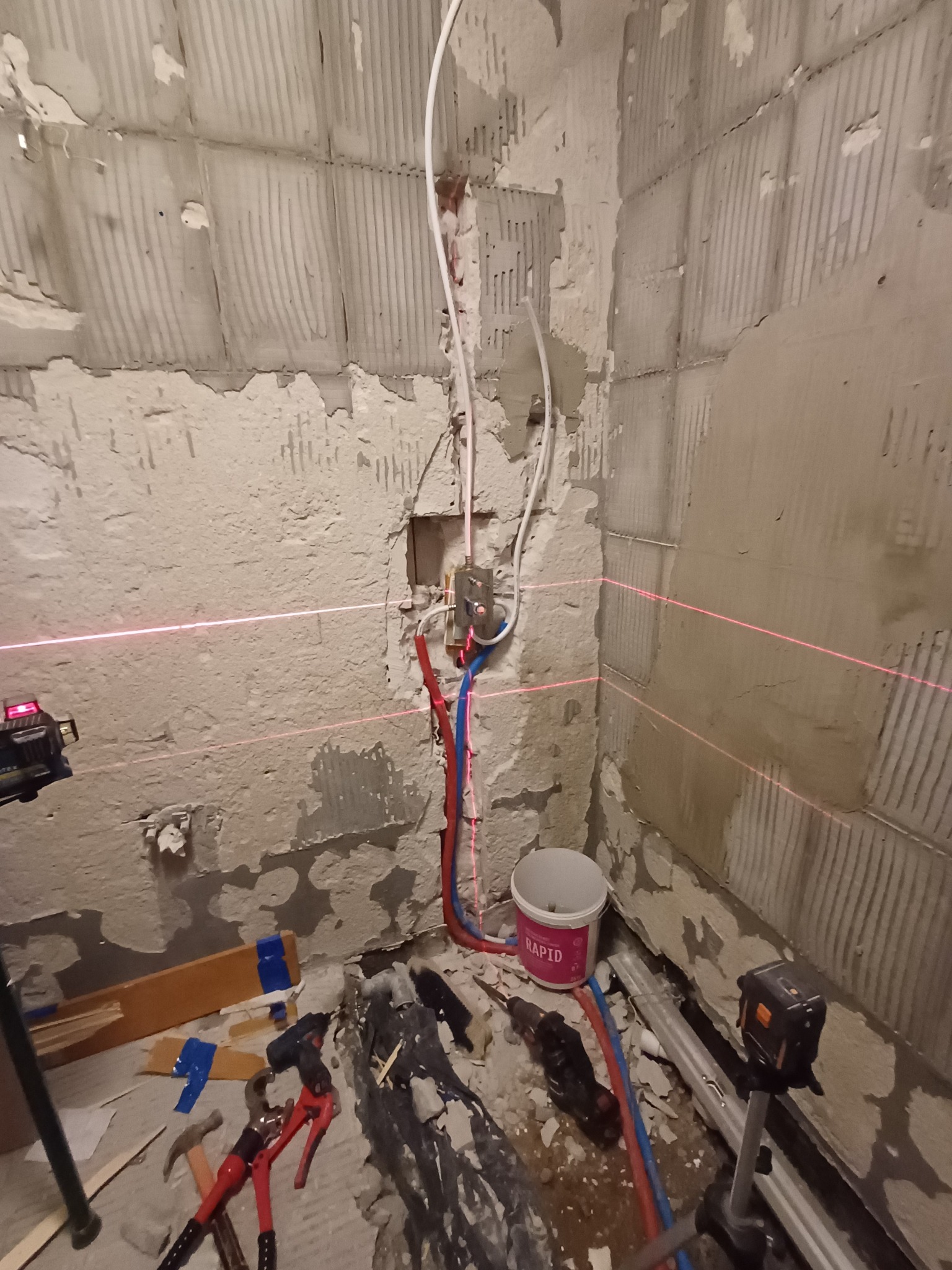 Przygotowanie do położenia płytek w łazience, widoczne gołe ściany, rury, poziomica laserowa, narzędzia oraz wiadro z farbą Rapid.