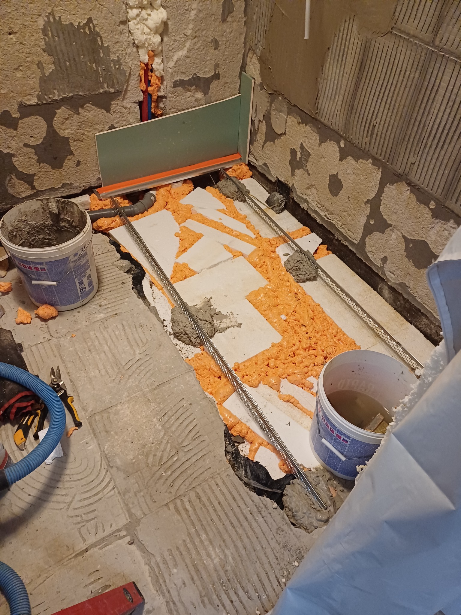 Przygotowanie podłoża pod prysznic: styropian, pianka montażowa, listwa odpływowa, rury odpływowe i wiadro z zaprawą w surowym wnętrzu łazienki.