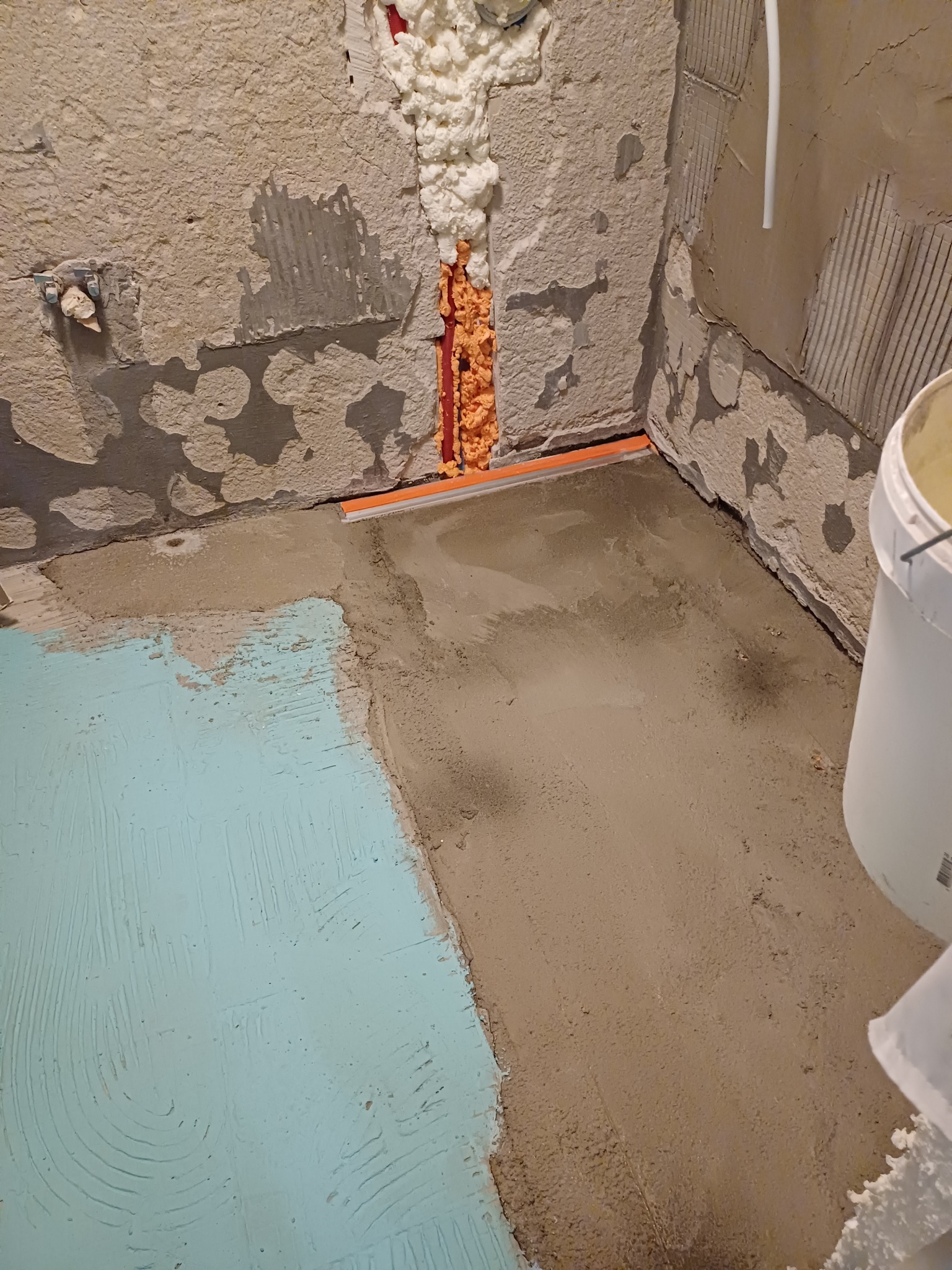 Przygotowanie podłoża w łazience: wylewka samopoziomująca częściowo pokryta niebieską hydroizolacją, ściany z resztkami starej glazury, wypełniona pianką montażową szczelina przy podłodze.