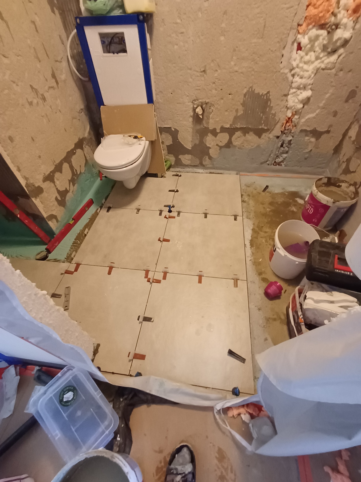 Niedokończona łazienka w trakcie układania jasnych, kwadratowych płytek podłogowych z użyciem krzyżyków dystansowych, częściowo otynkowane ściany z widoczną instalacją toalety podtynkowej i pianą...